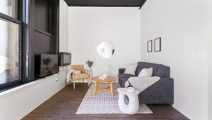 Un salon moderne avec un canapé gris, une chaise en bois, une petite table ronde et une table d’appoint blanche. Une grande fenêtre se trouve à gauche et un miroir est accroché au mur.
