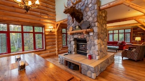 A log cabin interior with a stone fireplace, wooden beams, and large windows.