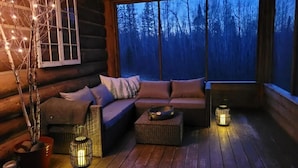 A cozy log cabin interior with a wooden deck, a sofa with cushions, a wicker ottoman, and a window with a view of a forest.