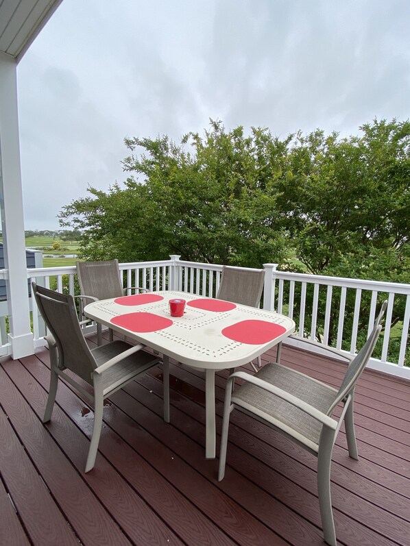 A covered outdoor patio with a round table and four chairs, a red cup on the table, and a view of trees and a golf course in the background. - Beaches, Golf, Pool and More (Rehoboth Beach)