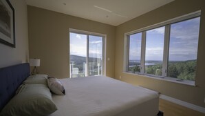 A bedroom with a large bed, two windows with views of a forest and a lake, and a lamp on a bedside table.
