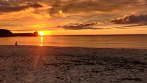 A beach with a sunset, calm sea, and a person sitting on the sand.