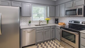 A modern kitchen with grey cabinetry, stainless steel appliances, a window with blinds, a sink, and a patterned floor. - Pet Friendly • BBQ • Patio • Desired Location (Charlotte)