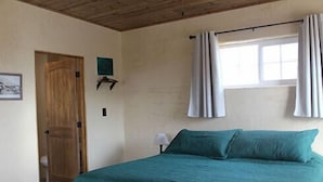 A bedroom with a bed, a wooden door, a window with curtains, and a wall-mounted light. - Big Bend Lodging at Ten Bits Ranch | Room #8 - The Courthouse (Terlingua)