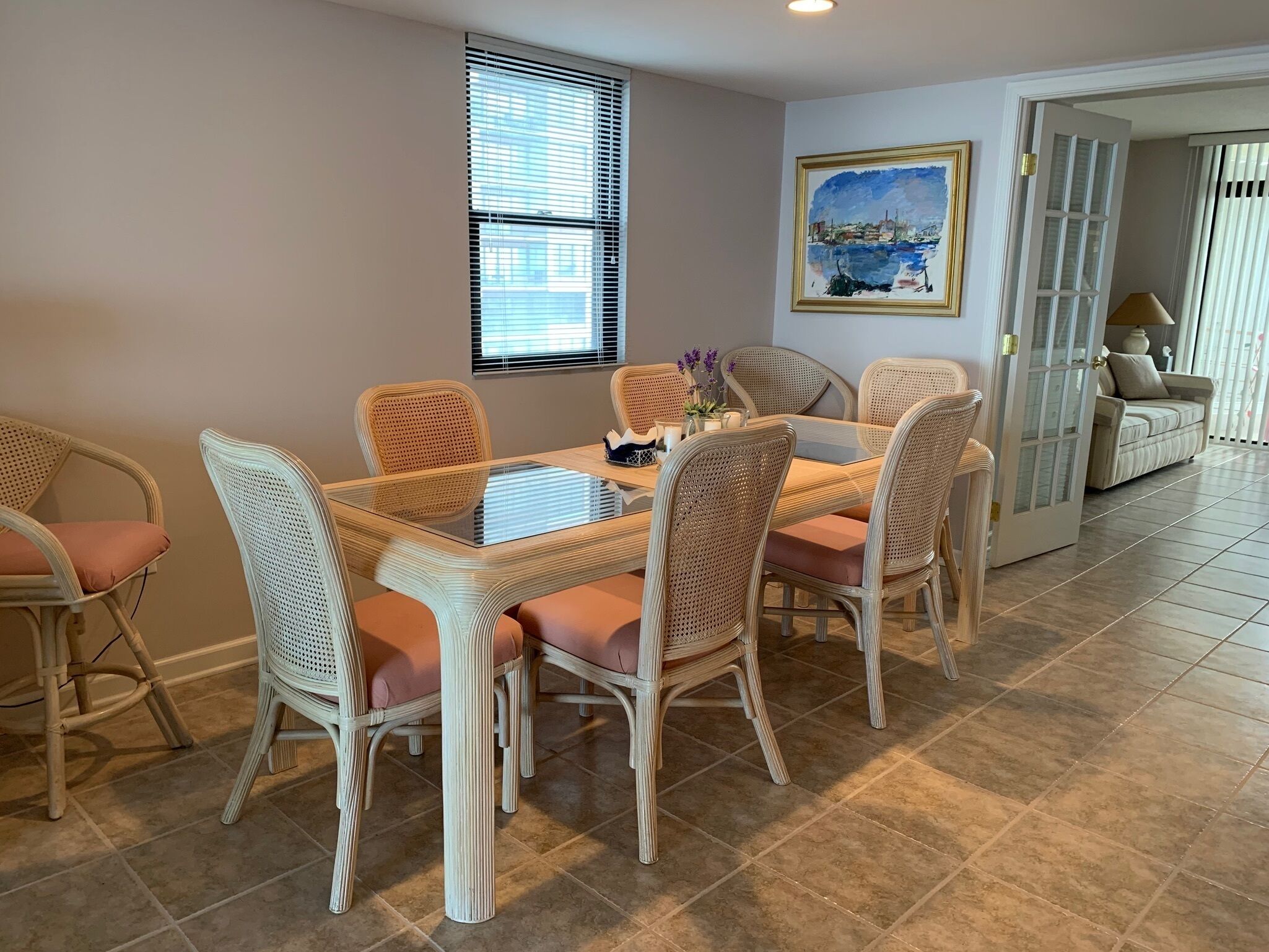 Un coin repas avec une table à plateau de verre entourée de chaises en osier, un tableau accroché au mur et une porte donnant sur une autre pièce.