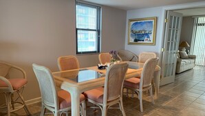 Un comedor con una mesa de vidrio rodeada de sillas de mimbre, un cuadro en la pared y una puerta que da a otra habitación.