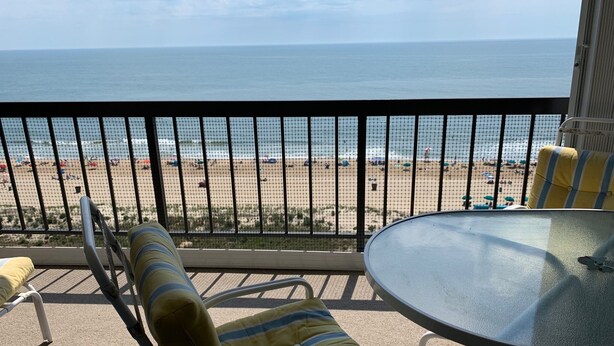 Un balcón con vista a la playa, una mesa redonda de vidrio y sillas a rayas.