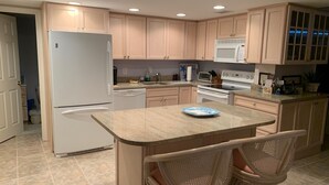Una cocina con armarios de madera clara, un refrigerador blanco, un microondas, una estufa y un lavamanos. Hay una pequeña isla con sillas y un plato.