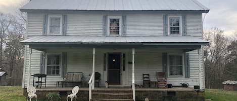 A two-story white house with a porch, a chimney, and a small front yard with a lawn and two chairs.