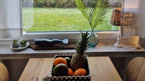 Un comedor con una mesa de madera, un frutero con piña, naranjas y pequeñas plantas en macetas, y vistas de un jardín y casas a través de una ventana.