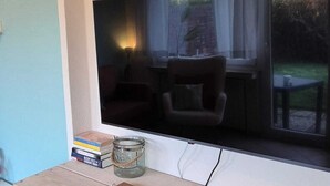 Una habitación con televisión montada en la pared, un escritorio de madera con un cuaderno, libros y un tarro. Debajo del escritorio hay dos cestas tejidas.