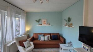 Una sala de estar con un sofá marrón, un sillón blanco, una mesita pequeña blanca, un televisor y un tapete estampado.