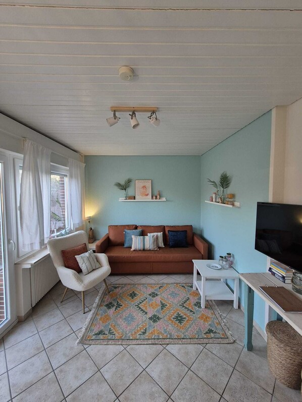 Un salon avec un canapé marron, un fauteuil blanc, une petite table blanche, un téléviseur et un tapis à motifs.