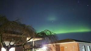 Una casa con un coche aparcado en la entrada, bajo un cielo nocturno con estrellas visibles y la aurora boreal.