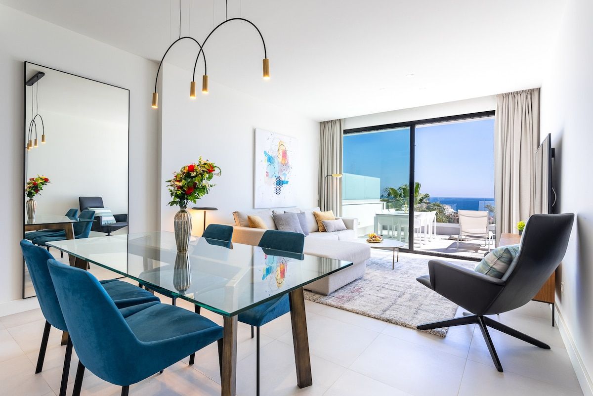 A modern living room with a glass dining table, blue chairs, a black armchair, and a large window offering a view of the ocean.