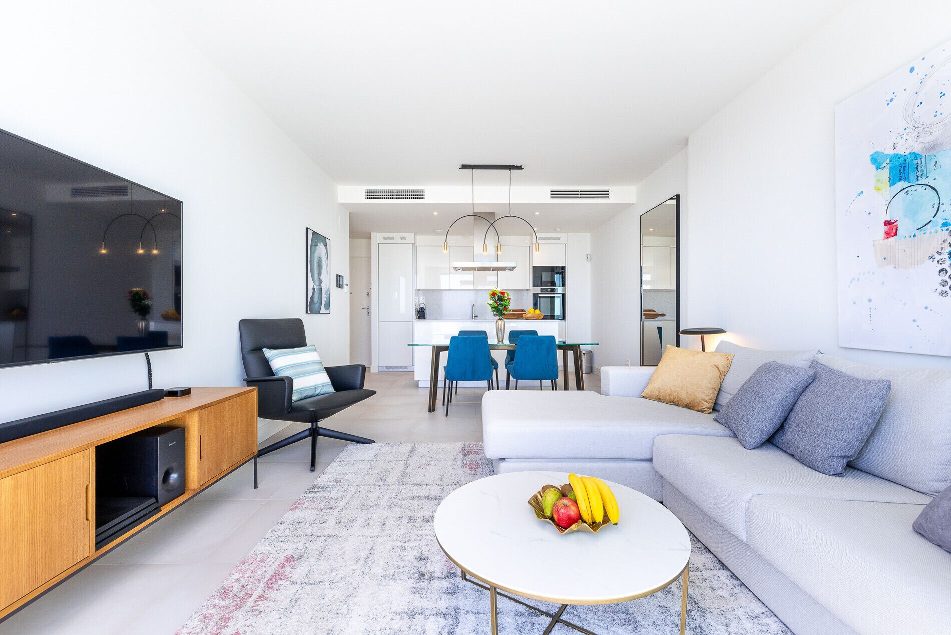A modern living room with a flat-screen TV, a sofa, a coffee table with fruit, and a dining area with chairs and a kitchen in the background.