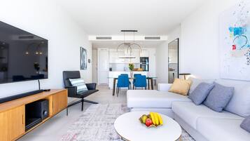 A modern living room with a flat-screen TV, a sofa, a coffee table with fruit, and a dining area with chairs and a kitchen in the background.