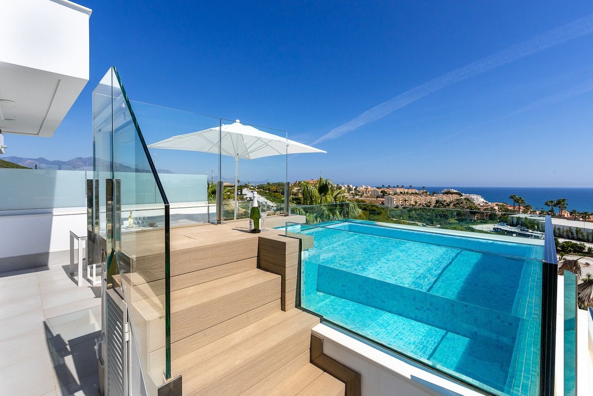 Una piscina en la azotea con barandillas de vidrio, terraza de madera y vista despejada de la costa.