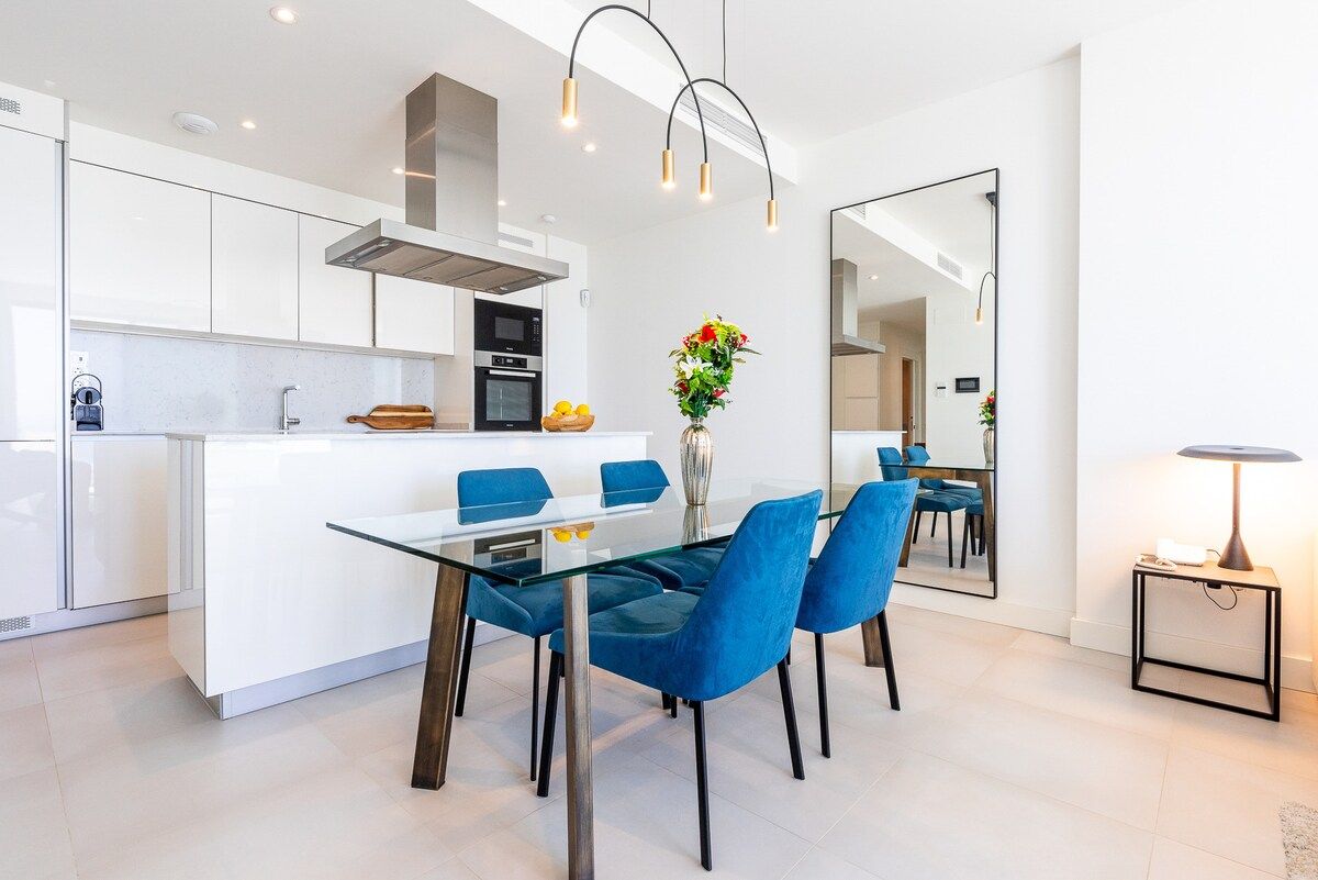 Una cocina moderna con armarios blancos, una isla central con asientos y un comedor con sillas azules.
