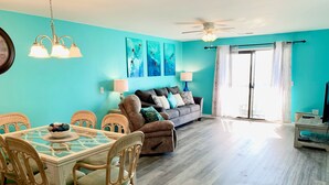 A living room with a teal wall, a wooden floor, a sofa with cushions, a dining table with chairs, and a ceiling fan.