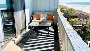 A balcony with a view of the beach, a sofa with cushions, a small table, and a red cup.