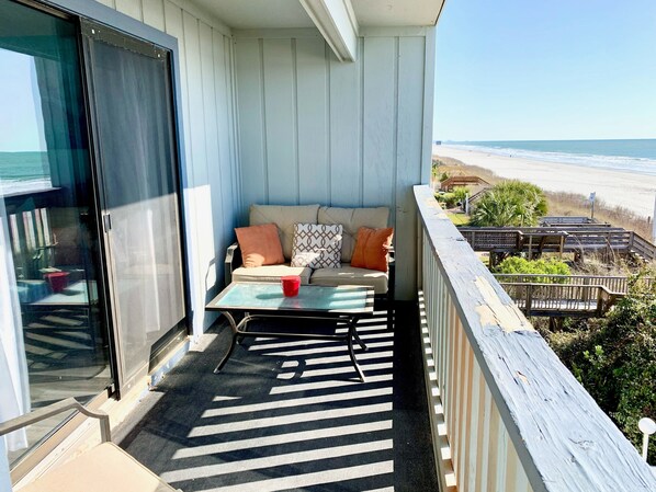 A balcony with a view of the beach, a sofa with cushions, a small table, and a red cup. - 2 Large Bedrooms, Double Oceanfront Balcony, Full size kitchen, Pool! (Surfside Beach)