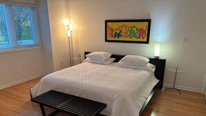 Un dormitorio con cama, mesitas de noche, un banco, una ventana con persianas y una obra de arte colgada en la pared.