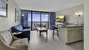 A modern living room with a sofa, a coffee table, and a dining area with a table and chairs. There is a kitchen area with a counter and cabinets. The room has large windows with a view of the sky and a balcony.