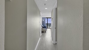 A corridor with a ceiling light, a door on the right, and a view of a balcony with a chair and a table.