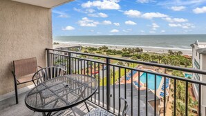 A balcony with a table and chairs overlooking a beach and a swimming pool.