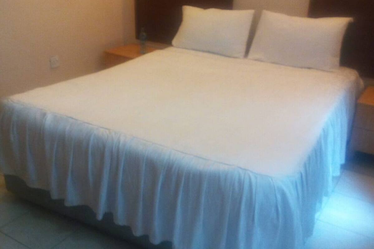 Uma cama com colcha e almofadas brancas, uma mesinha de cabeceira de madeira e uma tomada na parede.