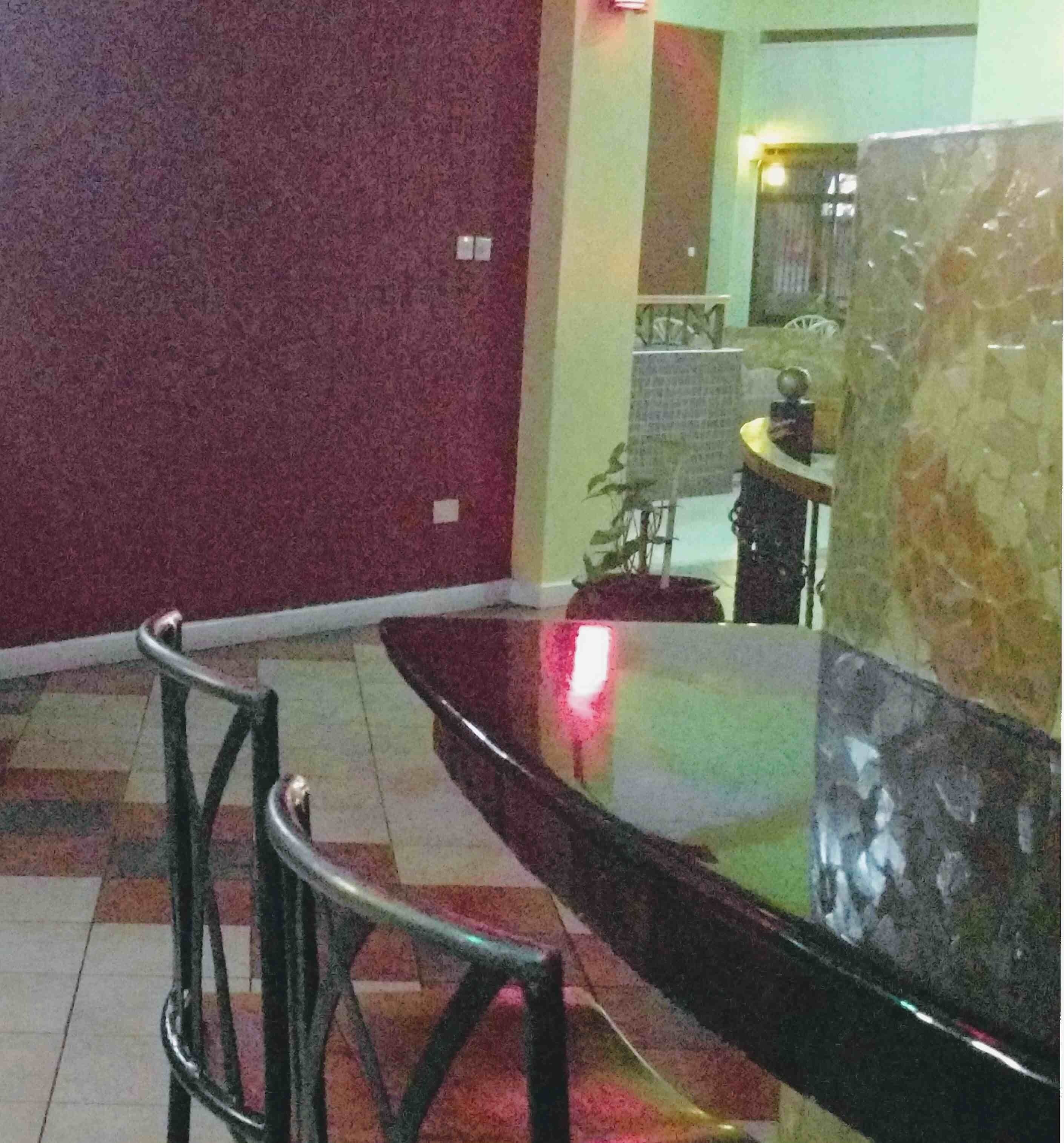 A dining area with a glass table and chairs, a red wall, and a kitchen area in the background.