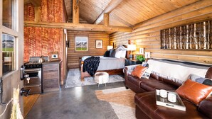 L’intérieur d’une cabane en rondins, avec un plafond en bois, un lit, un canapé, un petit coin cuisine et une grande fenêtre.