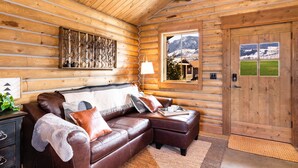L’intérieur d’une cabane en rondins, avec un canapé en cuir marron, un mur en bois orné d’une fenêtre donnant sur une montagne enneigée, et une porte en bois.