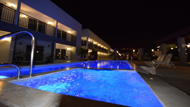 Una piscina con iluminación azul, junto a un edificio con balcones y sillones de descanso.