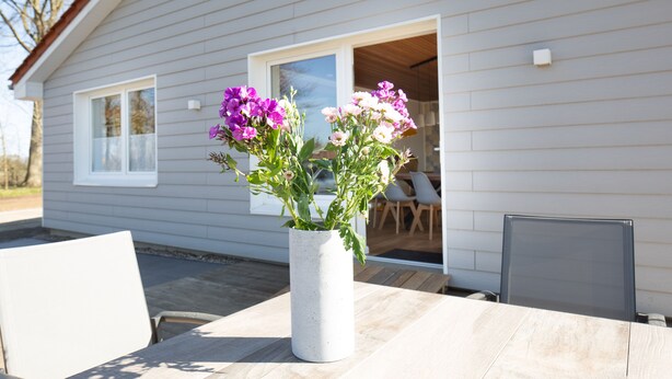 Een witte vaas met paarse en roze bloemen op een houten tafel voor een huis met een grijze gevel en een wit raam.