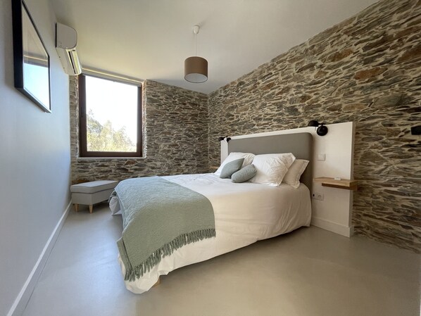 Un dormitorio con pared de piedra, una cama con almohadas, una mesita de noche y una ventana.