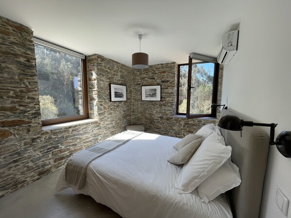 Un dormitorio con paredes de piedra, una cama con ropa de cama blanca, dos cuadros enmarcados y una ventana con vistas a árboles.