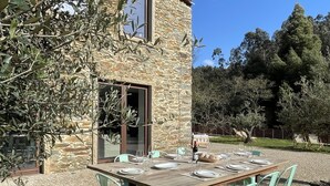An outdoor dining area with a stone wall, olive tree, and a view of the surrounding landscape.