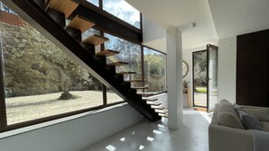 A modern interior with a staircase leading to a balcony, a sofa, and a view of a stone wall and greenery.