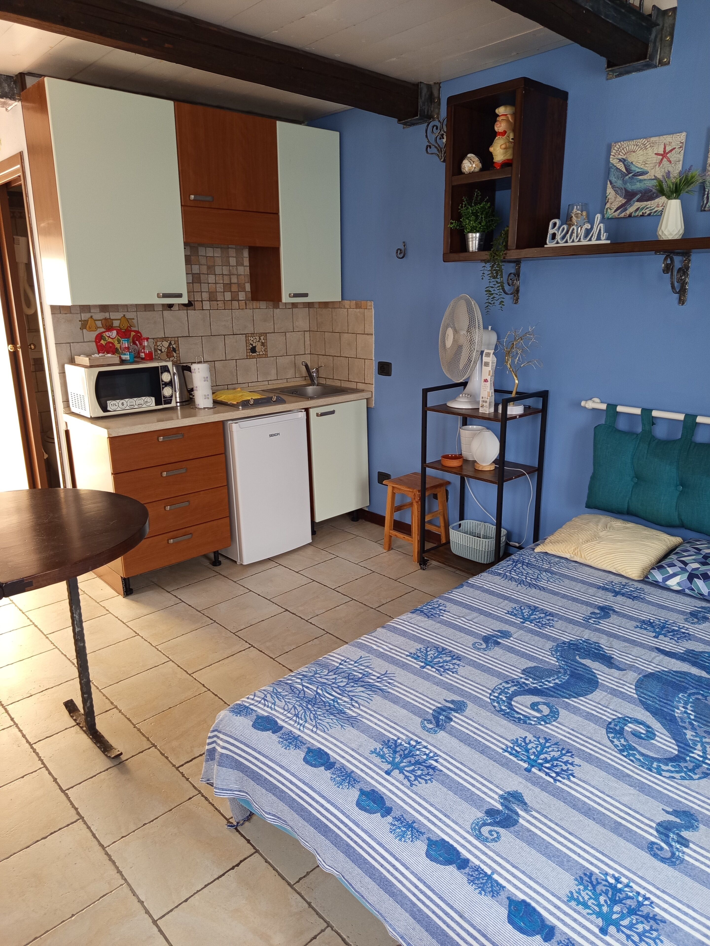 A compact room with a kitchenette, a bed with a blue and white patterned bedspread, a small wooden stool, and a shelf with decorative items.