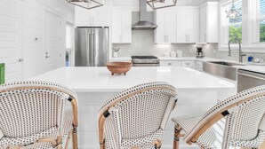 A modern kitchen with white cabinetry, a large island with a wooden bowl, and three wicker chairs. - Golf Cart｜4bed, 3.5 bath｜Luxury 30A Getaway! (Santa Rosa Beach)