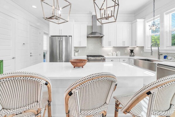 A modern kitchen with white cabinetry, a large island with a wooden bowl, and three wicker chairs. - Golf Cart｜4bed, 3.5 bath｜Luxury 30A Getaway! (Santa Rosa Beach)