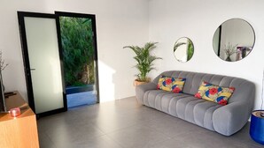 Un salon moderne avec un canapé gris, deux coussins colorés, une table d’appoint en bois, une plante en pot et deux miroirs muraux.