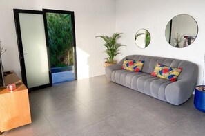 Een moderne woonkamer met een grijze bank, twee kleurrijke kussens, een houten bijzettafeltje, een potplant en twee spiegelwanden.