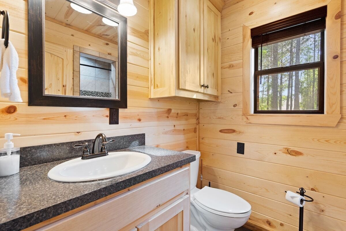 A wooden cabin bathroom with a granite countertop sink, a white toilet, and a window with a view of trees.