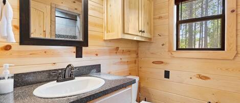 A wooden cabin bathroom with a granite countertop sink, a white toilet, and a window with a view of trees.