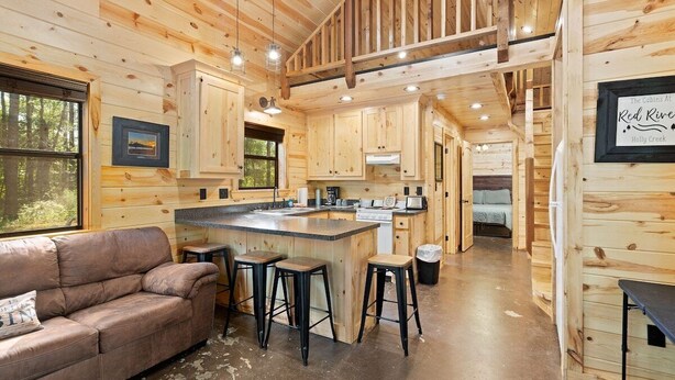 A cabin interior with wooden walls, a kitchenette, a dining area with stools, and a sofa. - Beautiful cabins with a pool in a peaceful setting (Broken Bow)