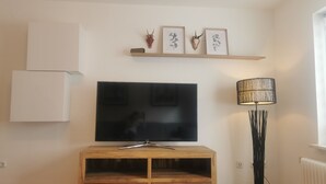 A living room with a flat-screen TV on a wooden TV stand, a floor lamp, and framed wall art above the TV. - Schwarzwaldmarie Ferienwohnung (löffingen)