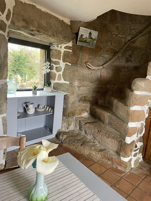 A stone-walled room with a rustic aesthetic, featuring a window with a view of greenery, a small shelf with decorative items, and a table with a vase of flowers.